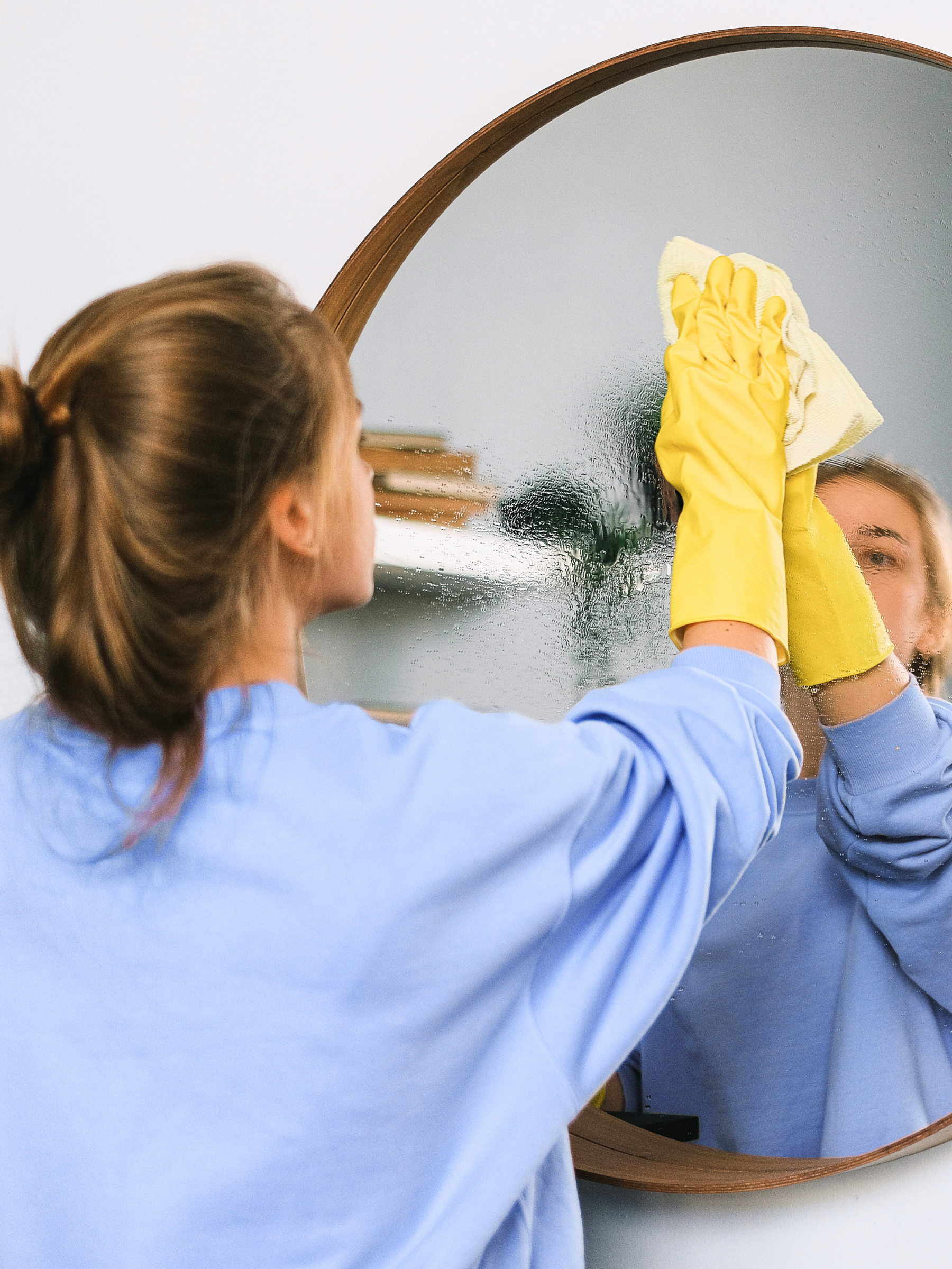 Henkilö puhdistaa peiliä pyöreämuotoisessa kehyksessä käyttäen keltaisia kumikäsineitä ja liinaa.