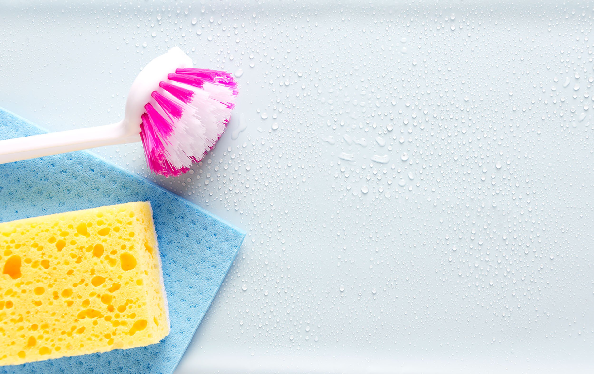 Kuvassa on pinkkivalkoinen tiskiharja, keltainen sieni ja sininen siivousliina kostean pinnan päällä.