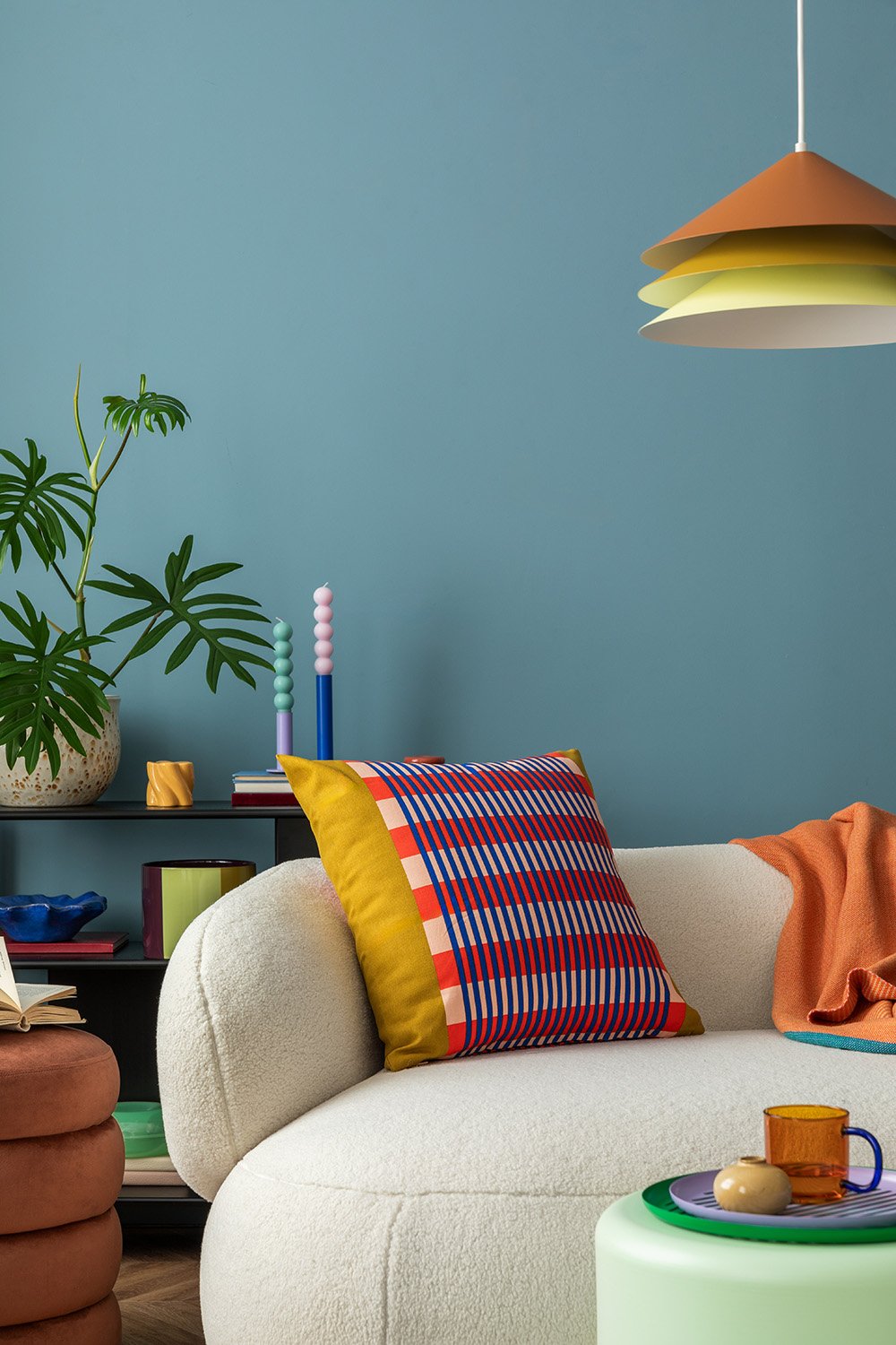 Värikäs sisustus, jossa on valkoinen sohva, ruudullinen tyyny, ruskea jakkara, oranssi peitto, vihreä pöytä, oranssi ja keltainen riippuvalaisin, seinähylly, maljakossa viherkasvi ja kynttilöitä vaaleansinisellä seinällä.