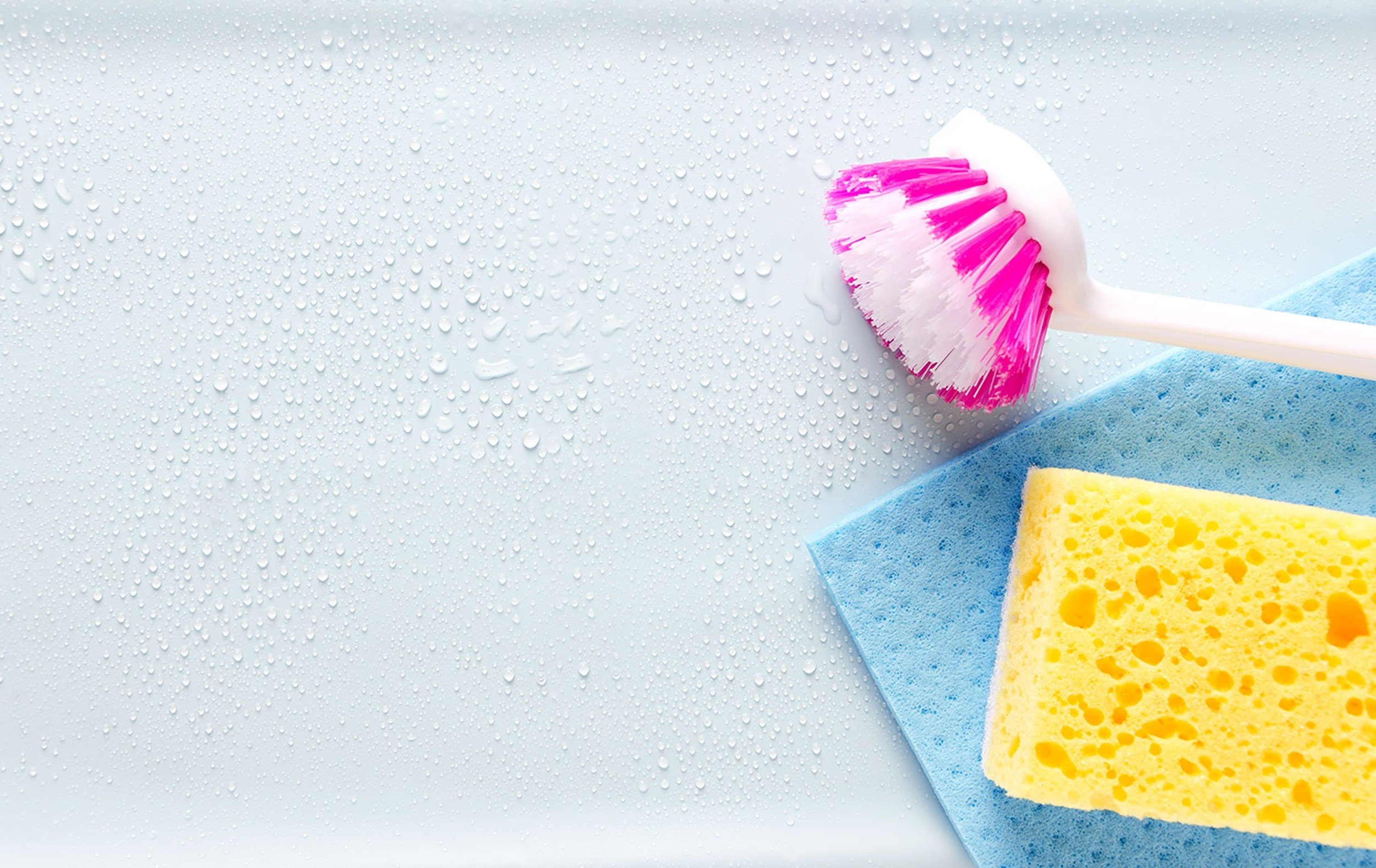 Keltainen sieni ja sininen puhdistusliina märällä pinnalla, vieressä valko-pinkki tiskiharja.